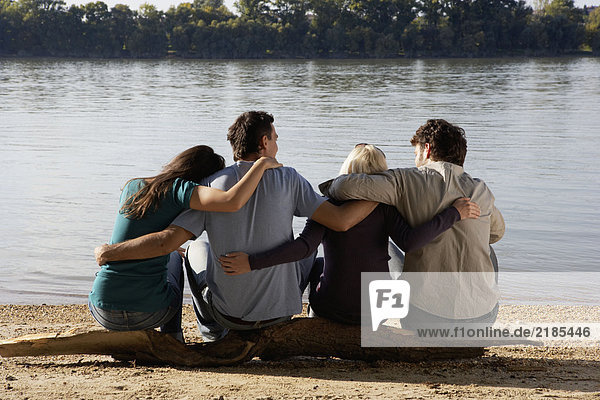 Four friends sitting on log by a lake with arms around each other.