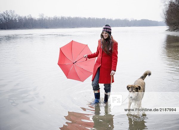 Junge Frau hält Regenschirm  watet im Fluss mit Hund  lächelt