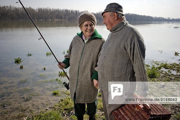 Senior couple carrying picnic basket and fishing rod by river  smiling