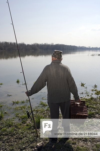 Senior man standing by river holding fishing rod  rear view
