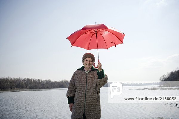 Seniorin am Fluss stehend mit rotem Regenschirm  lächelnd  Portrait