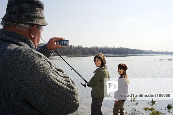 Großvater fotografiert Enkel (12-14) und Enkelin (10-12) beim Fischen im Fluss.
