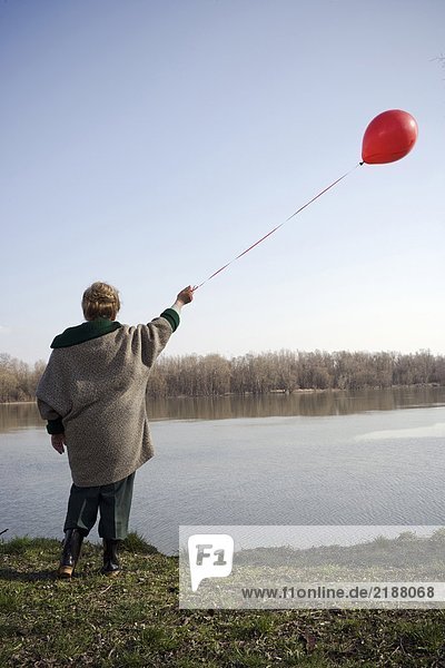 Senior woman standing by river holding red balloon  rear view