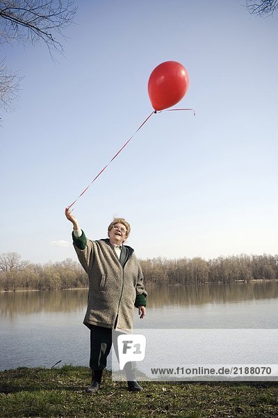 Senior woman standing by river holding red balloon  smiling