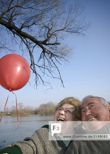 Senior couple by river holding red balloon  heads together  smiling