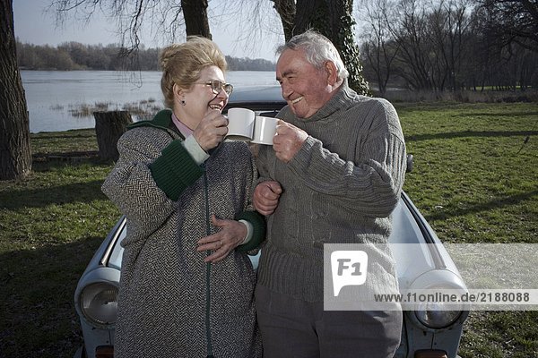 Senior couple standing by car toasting each other with cups  smiling