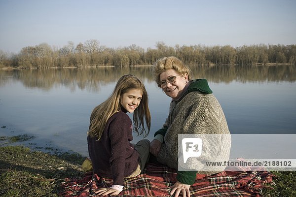 Grandmother and granddaughter (10-12) sitting on rug by river  smiling