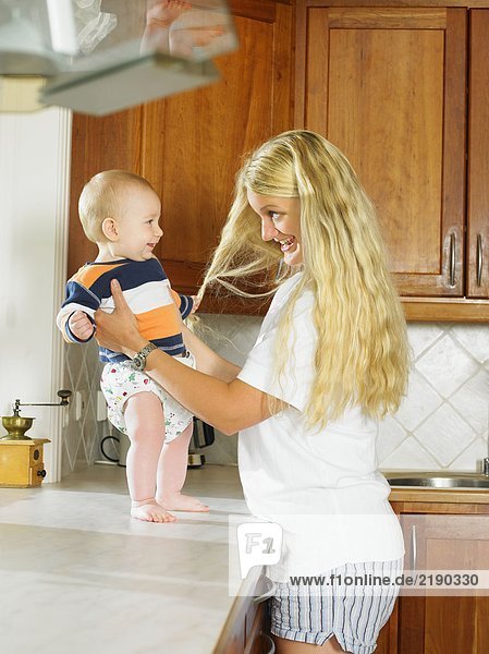 Frau hält Baby auf der Küchenzeile lächelnd hoch.