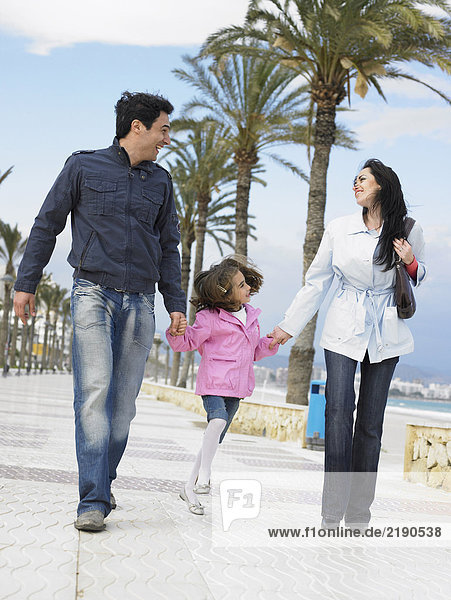 Mother and father walking along palm lined pavement by the sea with young daughter (6-8). Alicante  Spain.