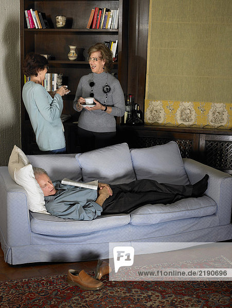 Älterer Mann schläft auf Sofa  zwei Frauen unterhalten sich im Hintergrund