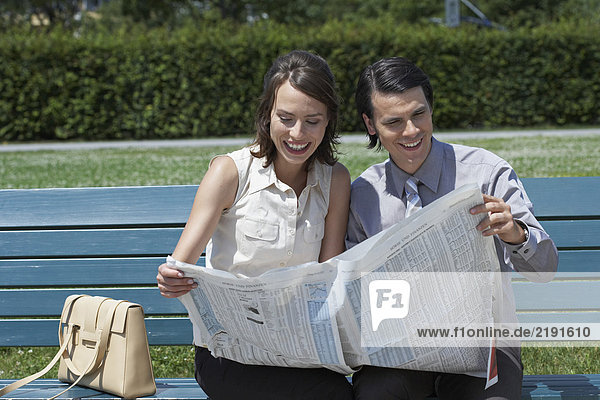 Geschäftsmann und Frau auf der Bank im Park beim Lesen einer Zeitung zusammen lächelnd.