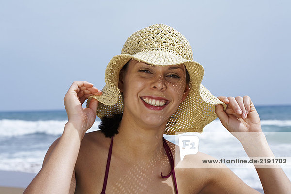 Junge Frau im Bikini am Strand mit Strohhut und Blick ins Meer im Hintergrund.