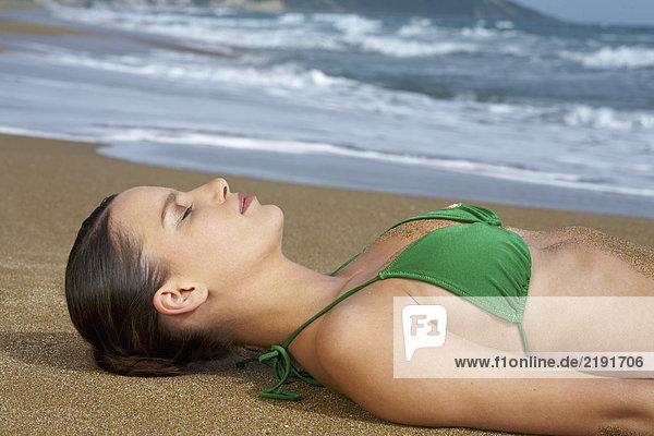 Junge Frau im Bikini am Strand liegt in Sand Augen geschlossen Meer im Hintergrund.