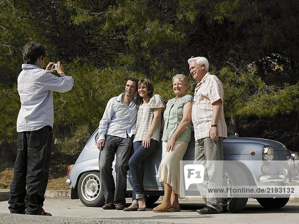 Multi-generational family standing beside car boy (8-10) taking photo