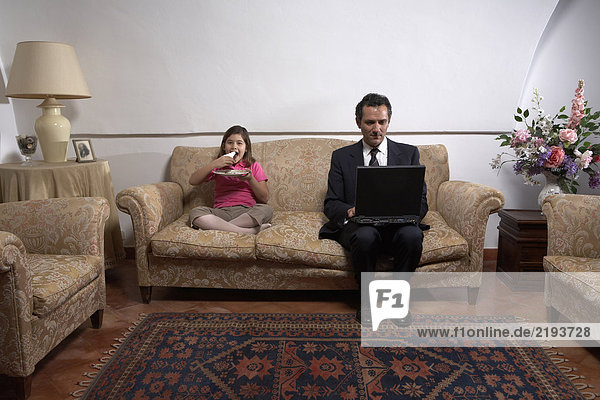 Man using laptop with daughter (6-8) on sofa eating cake