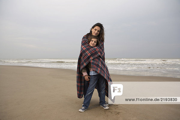 Mother embracing daughter (9-11) in blanket on beach  portrait