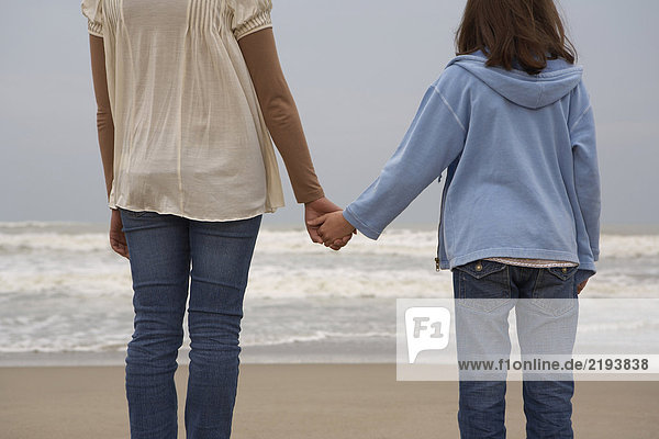 Mother and daughter (9-11) holding hands on beach  rear view