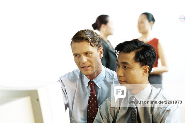 Two businessmen looking at desktop computer together  women standing in background