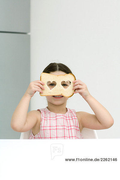 Kleines Mädchen hält Brot mit herzförmigen Ausschnitten hoch und schaut durch die Löcher in die Kamera.
