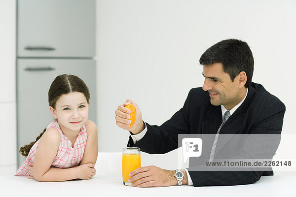 Vater und Tochter sitzen zusammen am Tisch  Mann drückt Orangensaft ins Glas  Mädchen lächelt in die Kamera