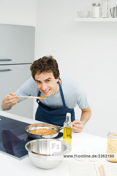 Mann kocht in der Küche  bückt sich  um Sauce zu schmecken  lächelt die Kamera an.