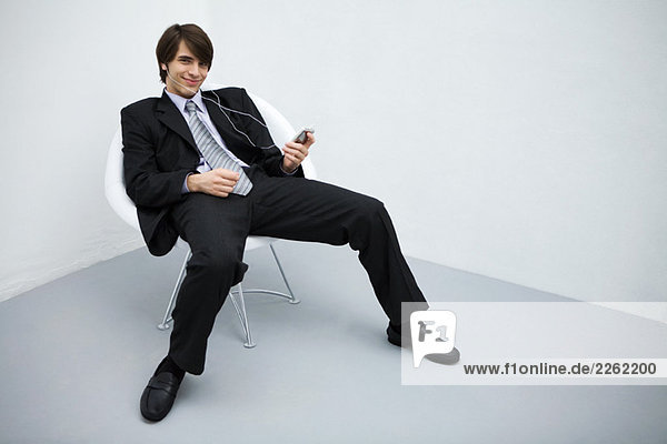 Young man in suit leaning back in chair  listening to mp3 player  smiling at camera  full length