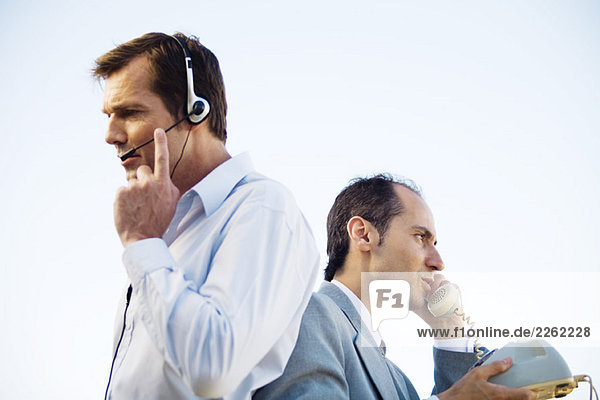 Two businessmen back to back  one using headset  the other using landline phone  side view