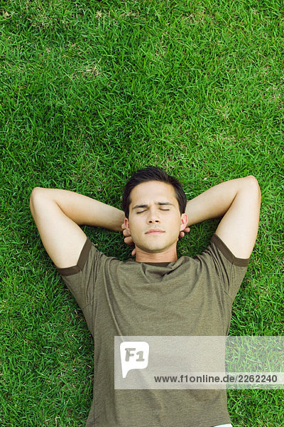 Young man lying on grass with hands behind head  eyes closed  high angle view