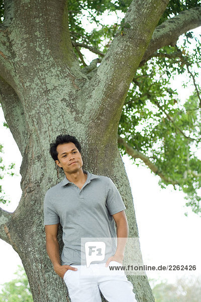 Mann an Baum gelehnt mit Händen in den Taschen  lächelnd