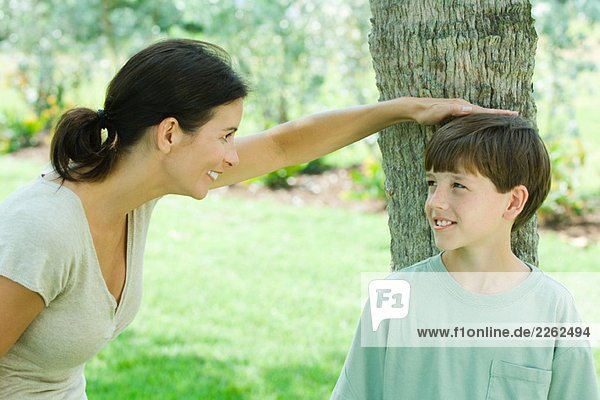 Junge lehnt an Baumstamm  seine Mutter legt ihre Hand auf seinen Kopf  beide lächelnd