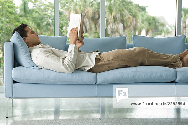 Young man lying on couch reading book  side view