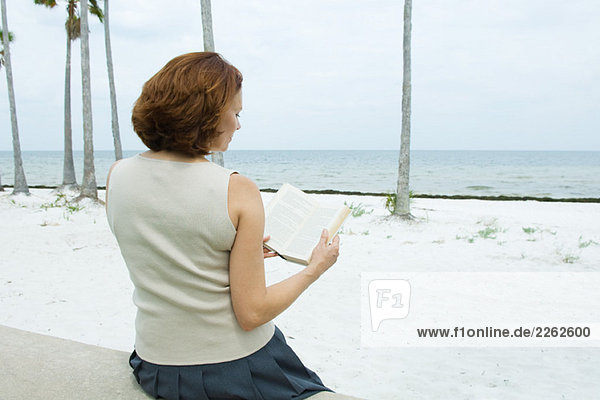 Frau sitzt am Strand und liest Buch  Rückansicht