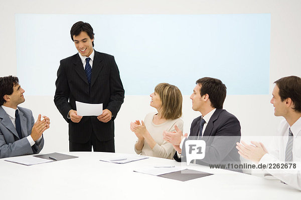 Businessman standing  holding document  seated colleagues clapping  smiling