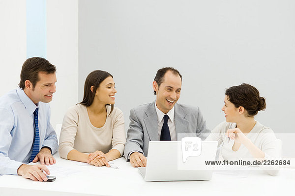 Gruppe von männlichen und weiblichen Geschäftspartnern  die zusammen am Tisch mit einem Laptop sitzen und lächeln.