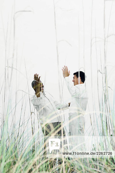 Zwei Männer grüßen sich draußen  die Hände erhoben  durch hohes Gras gesehen.