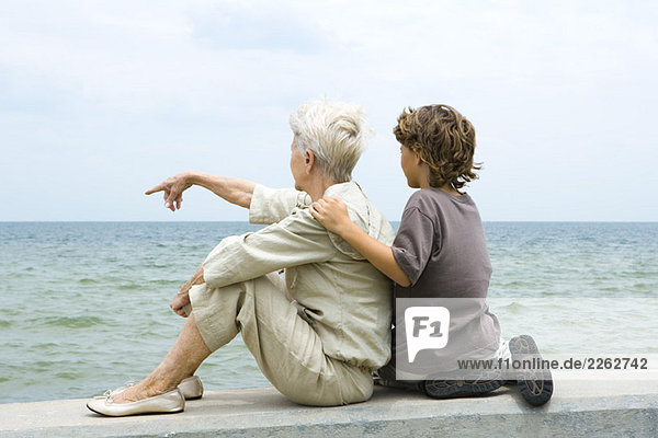 Großmutter und Enkel sitzen zusammen am Meer  Frau zeigt  Rückansicht
