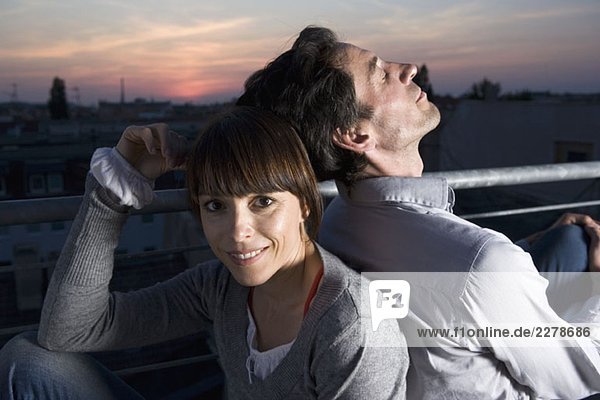Ein Paar  das nachts Rücken an Rücken auf einer Dachterrasse sitzt.
