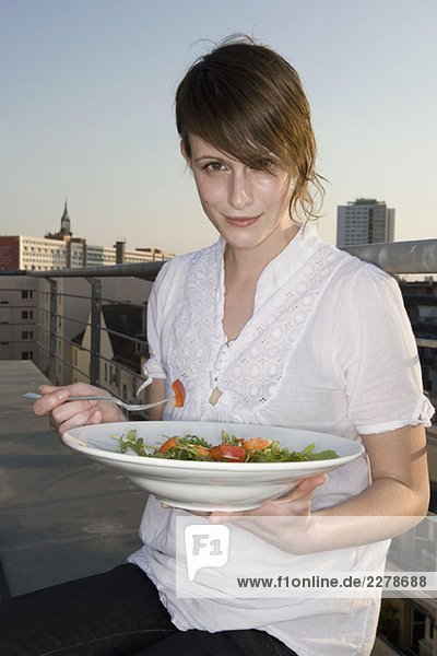 Eine junge Frau sitzt auf einer Dachterrasse und hält einen Teller Salat.