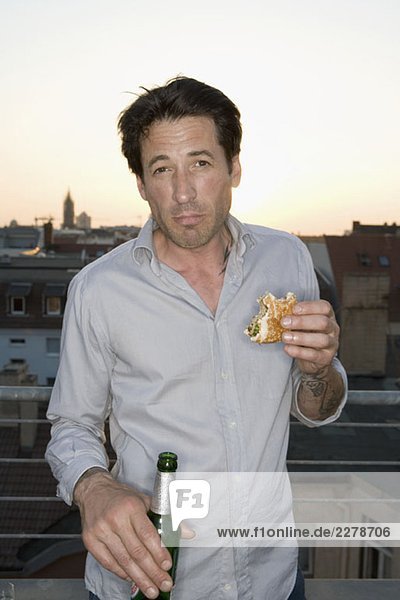 Ein Mann isst ein Sandwich und trinkt in der Abenddämmerung ein Bier auf der Terrasse.