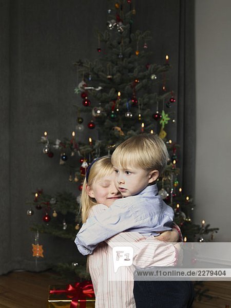 Ein Bruder und eine Schwester umarmen sich vor einem Weihnachtsbaum.