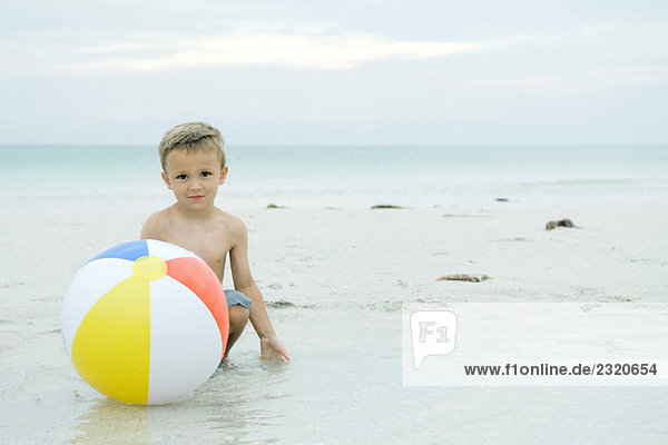 Kleiner Junge kauert hinter dem Ball am Strand und lächelt in die Kamera.
