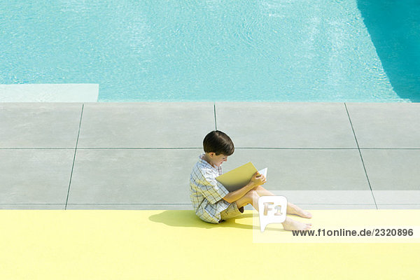 Boy sitting next to swimming pool  reading book  high angle view