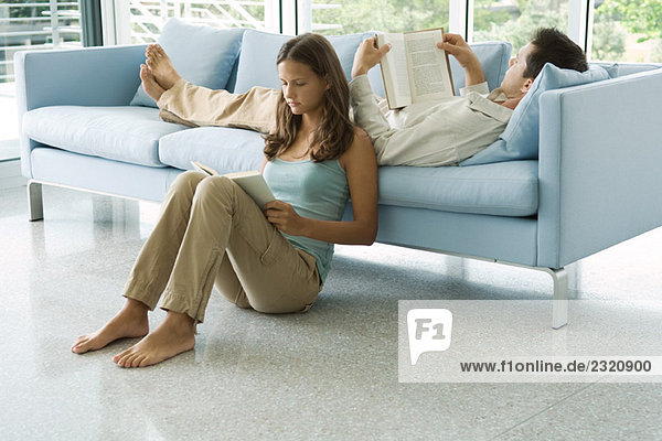 Young couple reading books together  male lying on sofa  female sitting on floor