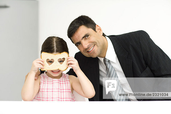 Vater und Tochter sitzen Seite an Seite  Mädchen schaut durch eine Scheibe Brot mit herzförmigen Löchern  beide lächeln in die Kamera.