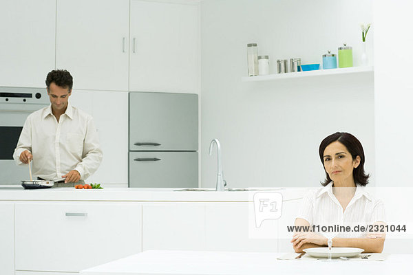 Woman sitting at table with arms folded  looking at camera  husband preparing meal in the background
