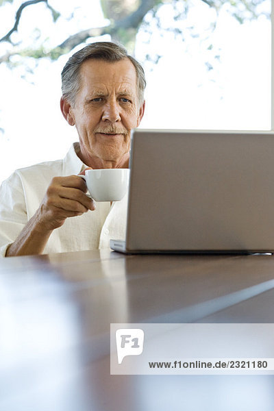 Senior Mann sitzt am Tisch  schaut auf den Laptop und hält eine Kaffeetasse.