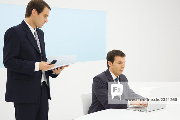 Two businessmen in office looking down at laptop computer together  one holding cell phone