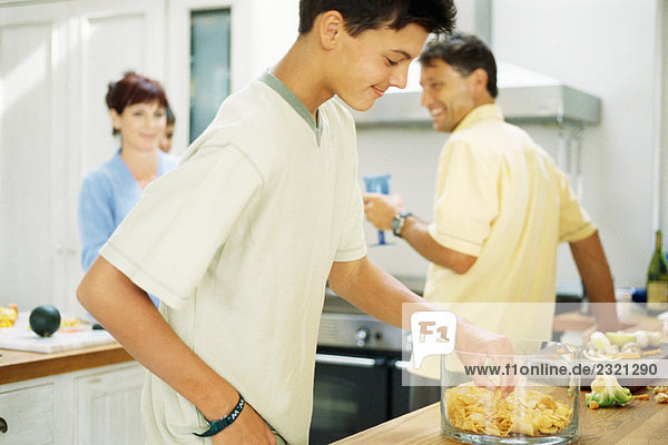 Teenager-Junge steht in der Küche  isst Pommes frites aus der Schüssel  Familie bereitet Essen im Hintergrund zu.