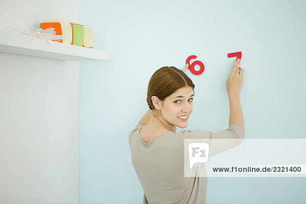 Frau arrangiert dekorative Nummern an der Wand  lächelt über die Schulter vor der Kamera