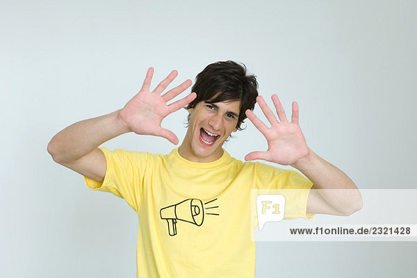 Junger Mann im T-Shirt mit Megaphon-Grafik  lächelnd vor der Kamera  Mund offen  Hände erhoben